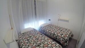 A small, clean room with two single beds, a window with curtains, and a bedside table with a lamp. - Beachfront apartment (La Cala del Moral)