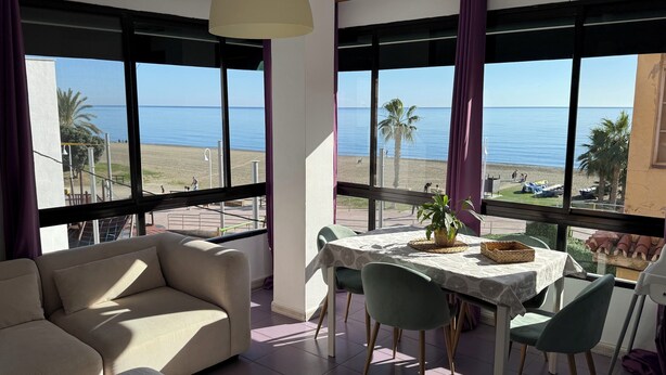 A room with a sofa, dining table, chairs, and large windows with a view of the beach and ocean. - Beachfront apartment (La Cala del Moral)
