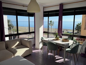 A room with a sofa, dining table, chairs, and large windows with a view of the beach and ocean.