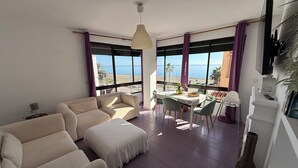 A living room with a sofa, a dining table with chairs, a TV mounted on the wall, and large windows with a view of the sea. - Beachfront apartment (La Cala del Moral)