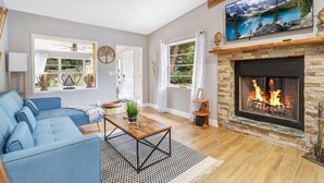 Ein Wohnzimmer mit einem blauen Sofa, einem Kamin, einem darüber montierten Fernseher und einem großen Fenster mit Bergblick.
