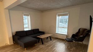 A spacious living room with a sectional sofa, a small coffee table, and a black armchair. There are two windows providing natural light.