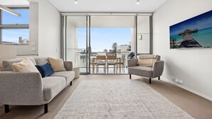 En moderne stue med en grå sofa, en mindre lænestol og et sofabord. Der er et stort vindue med udsigt til en balkon og et tropisk resort i baggrunden.