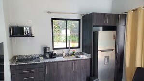 A modern kitchen with dark cabinetry, a stainless steel refrigerator, a coffee maker, and a window with a view of greenery. - One Life Villas (Tamarindo)