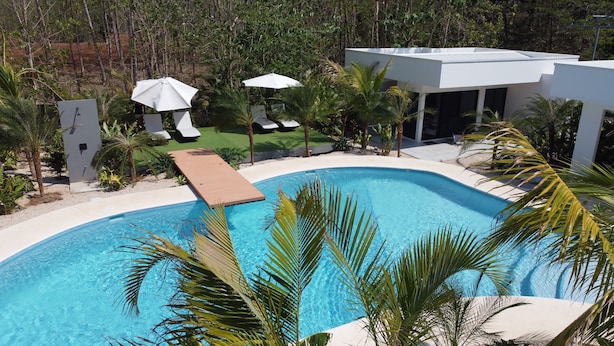 A swimming pool with a wooden deck, surrounded by palm trees and lounge chairs under white umbrellas. - One Life Villas (Tamarindo)
