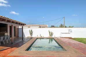 A swimming pool with a wooden deck, a covered patio area, and cacti against a white wall. - Villa 'Fatema' with Private Pool, Wi-Fi, and Air Conditioning (Conil de la Frontera)