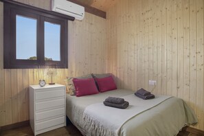 A bedroom with a wooden wall, a bed with a white bedspread and two pillows, a bedside table with a lamp, and an air conditioning unit above a window. - Villa 'Fatema' with Private Pool, Wi-Fi, and Air Conditioning (Conil de la Frontera)