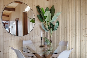 Uma sala de jantar com mesa de vidro, cadeiras brancas, uma planta em vaso grande e um espelho na parede.