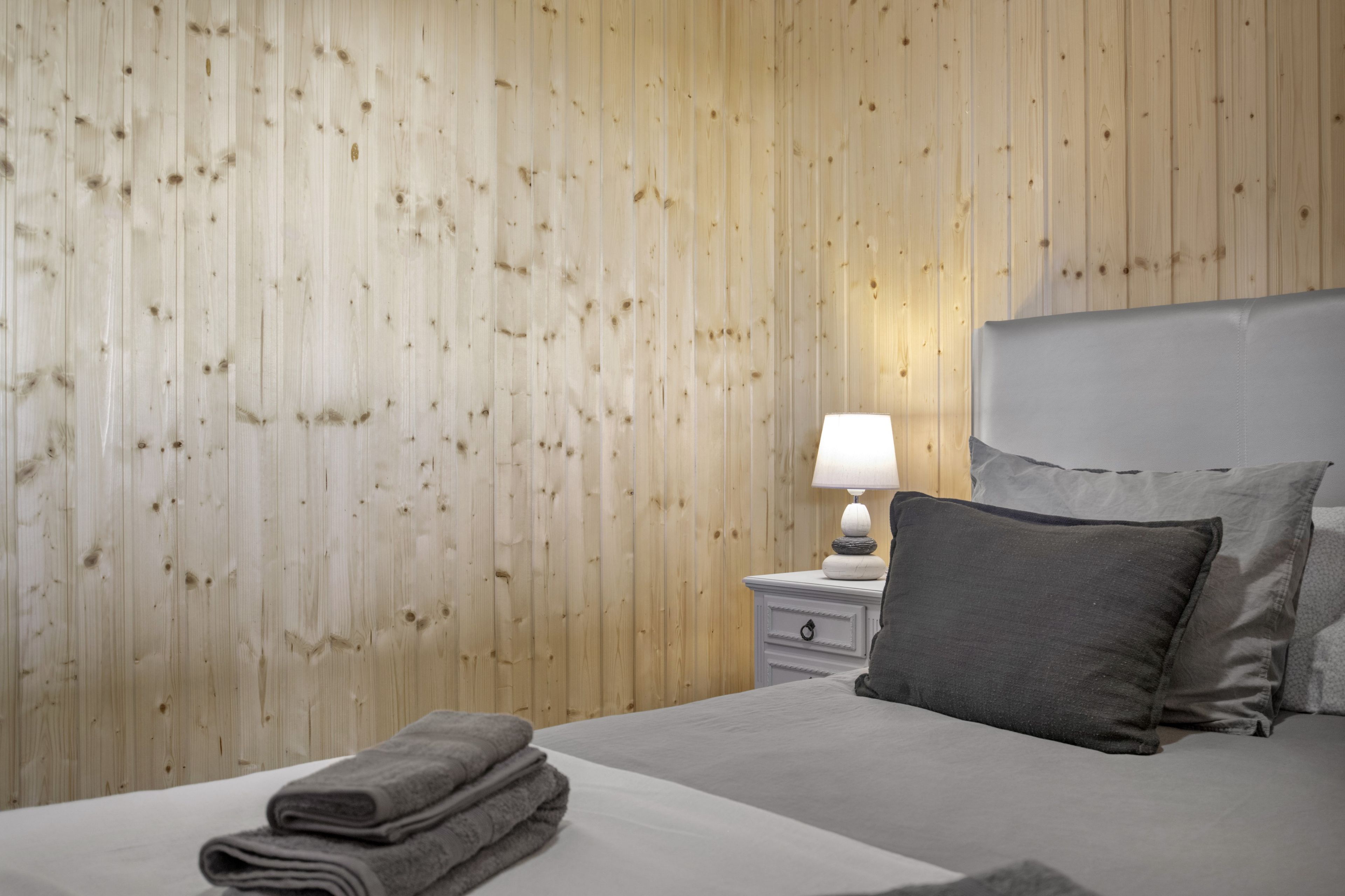 Une chambre d’hôtel avec un mur en bois, un lit avec une literie grise, une table de chevet avec une lampe et une serviette grise pliée.