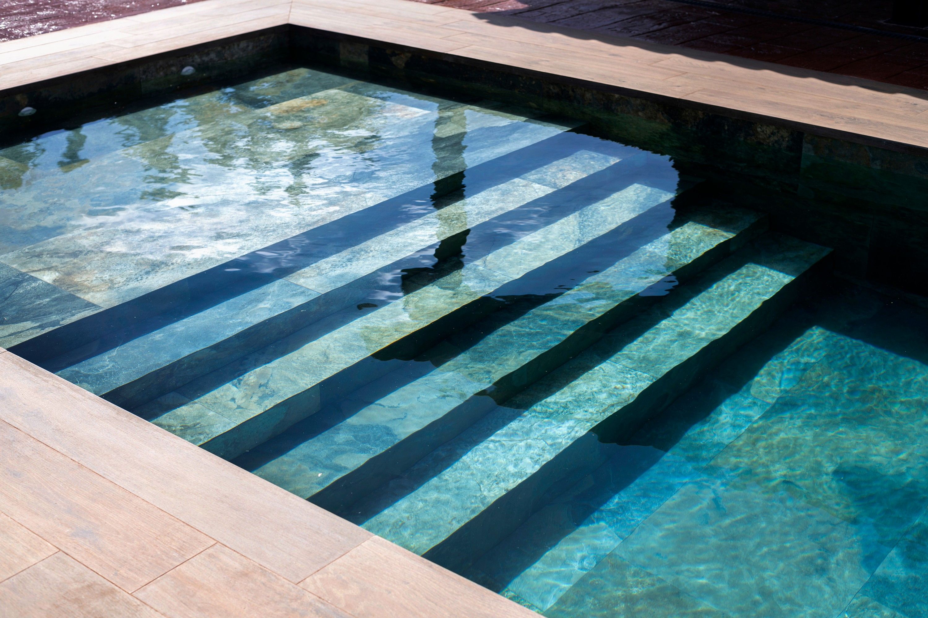 Una piscina rectangular y cristalina, con borde de piedra y un solárium con baldosas alrededor.