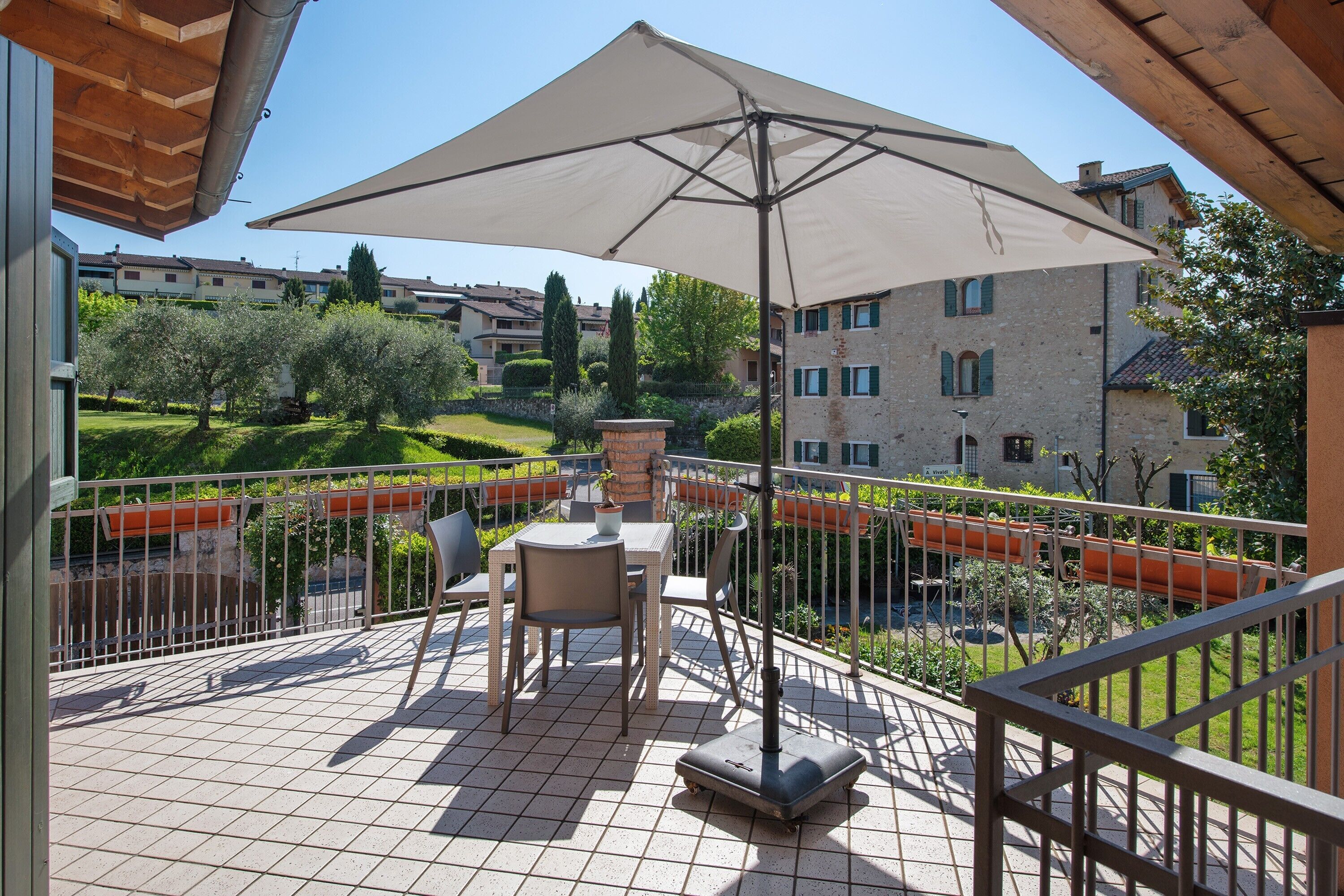 Een balkon met een tafel en stoelen, een grote parasol en uitzicht op een tuin en gebouwen op de achtergrond.