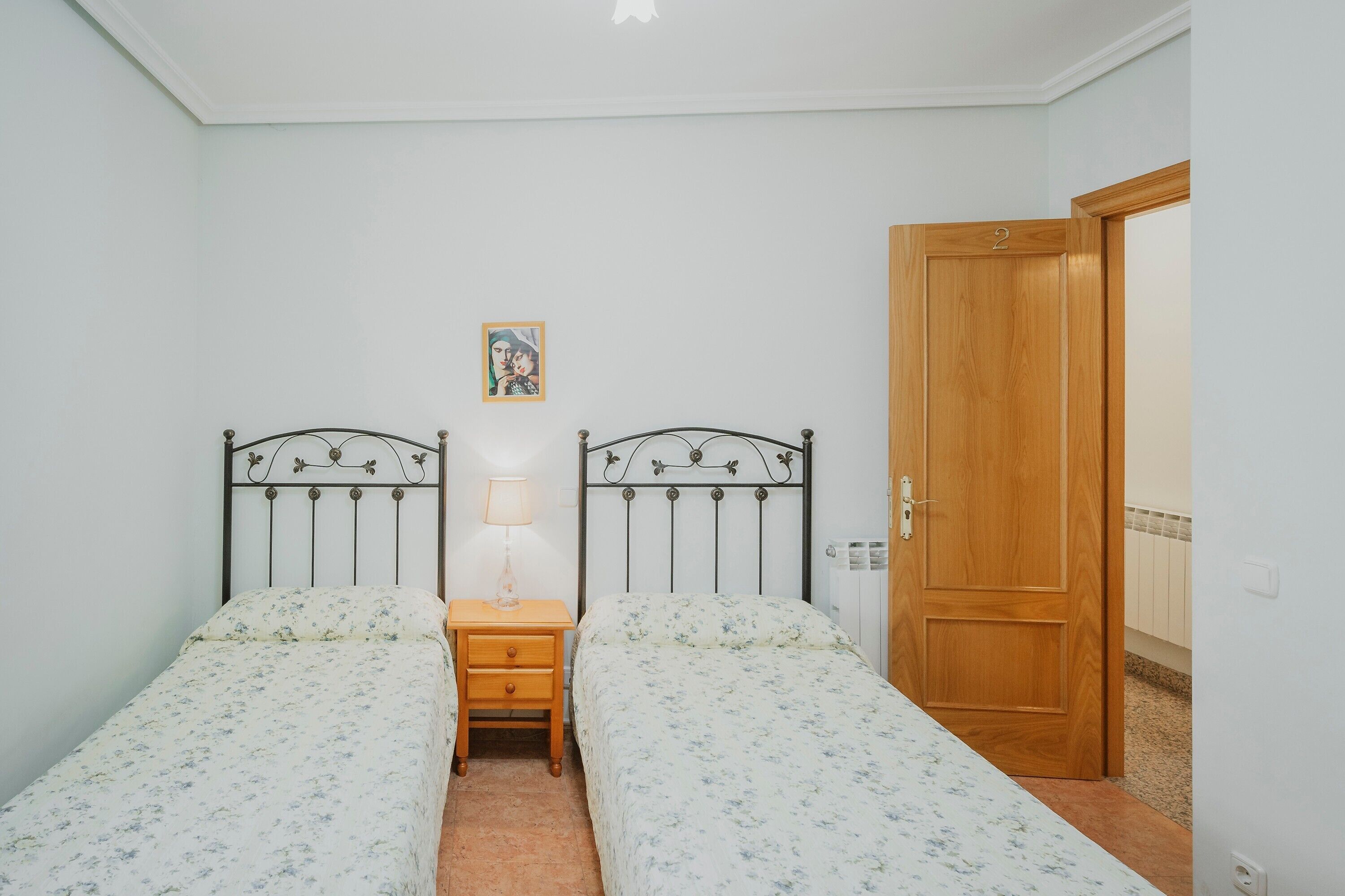 A room with two single beds, a bedside table with a lamp, a wooden door, and a small framed picture on the wall.