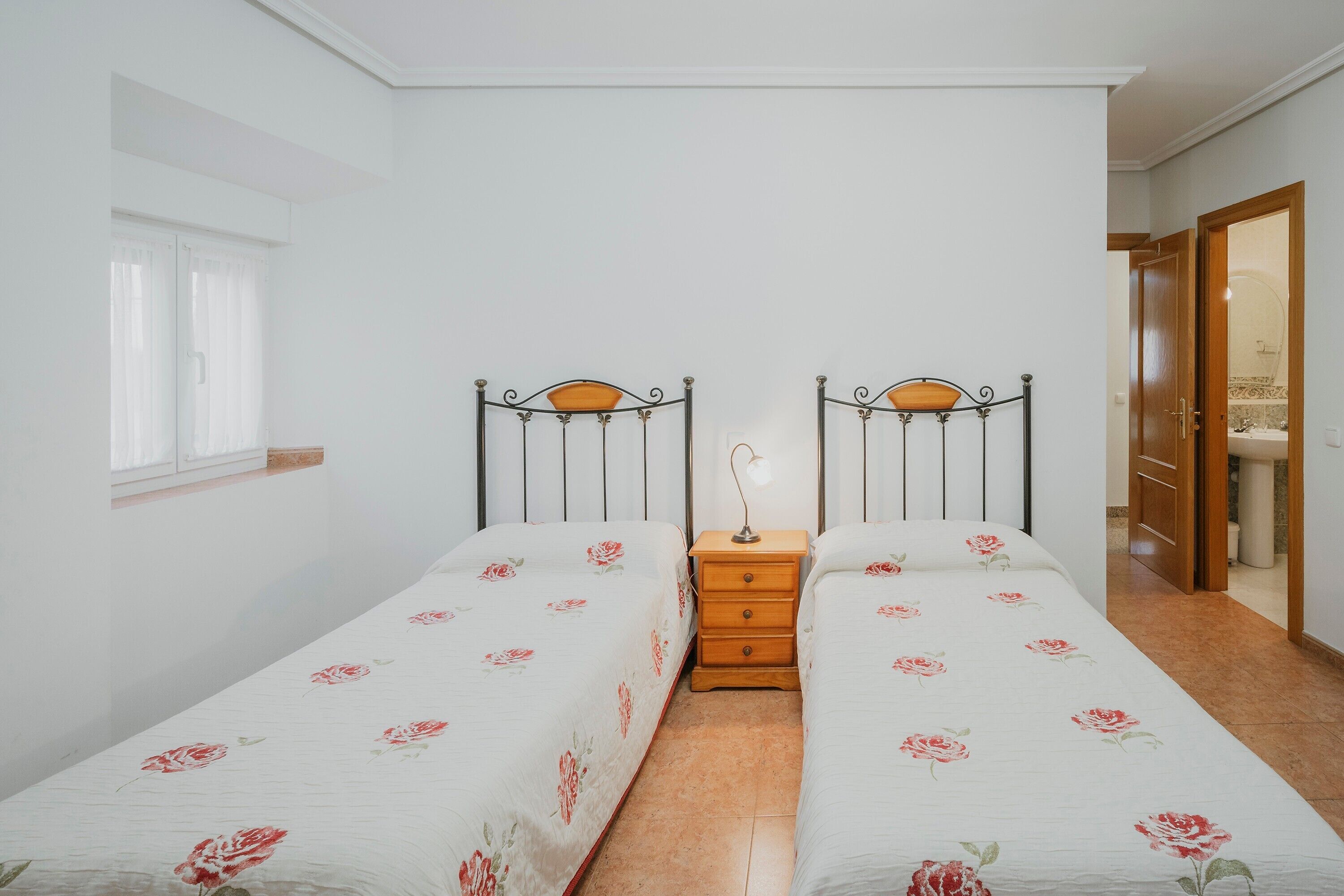 Une chambre avec deux lits simples, une table de chevet, une fenêtre avec des rideaux et une porte donnant sur une salle de bain.