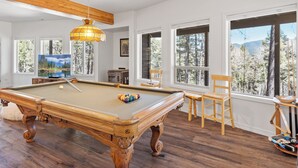 A pool table with a wooden frame and a hanging light fixture. There are windows with a view of trees and mountains.