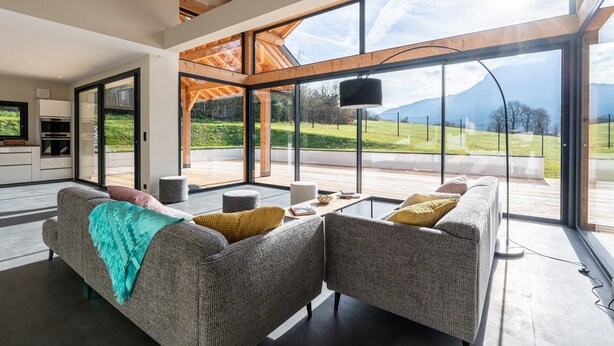 Una sala de estar moderna con sofá, reposapiés y mesa de centro, ventanas amplias con vista a la montaña, y una zona de cocina.