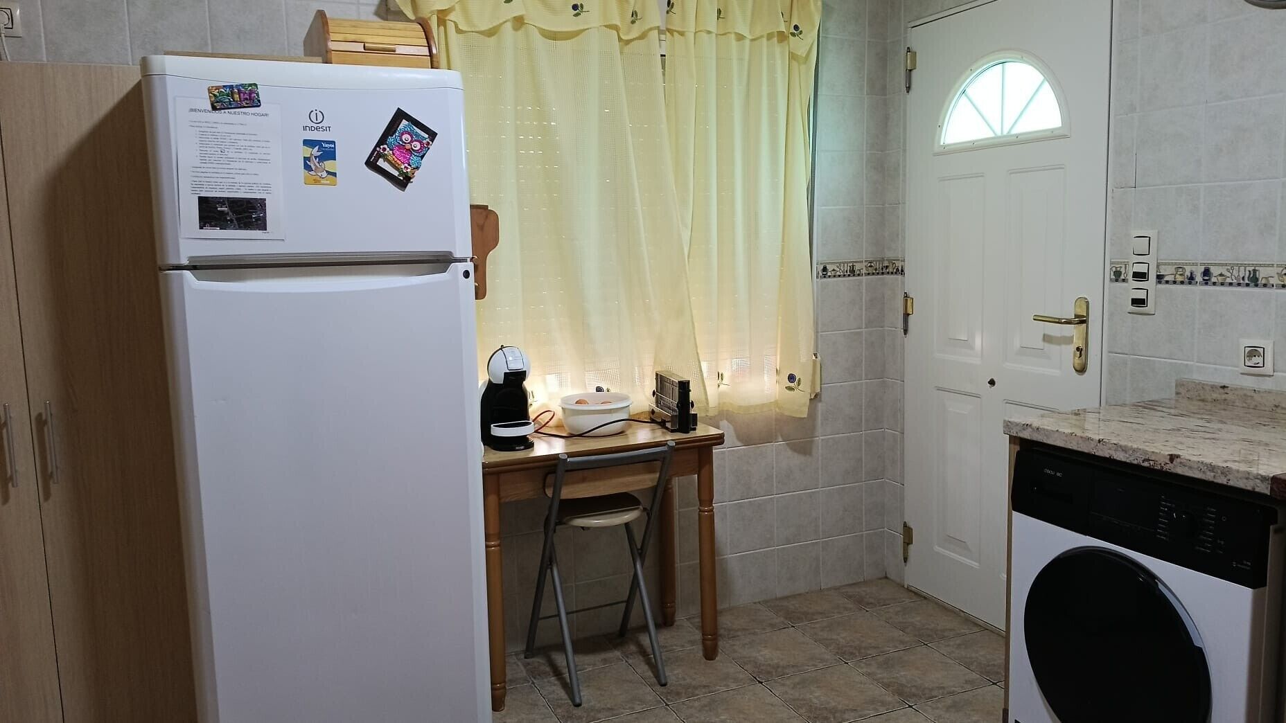 Cocina compacta con refrigerador, lavadora, una mesita con una silla y una ventana con cortina.