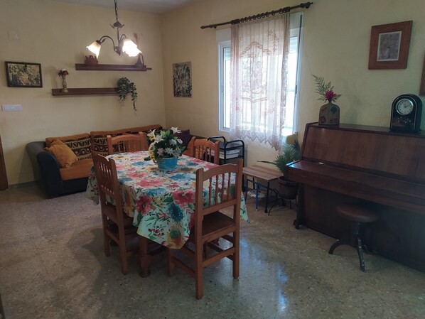 A dining area with a floral tablecloth, wooden chairs, a piano, and a sofa.