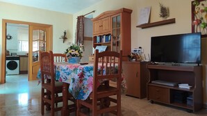 Ein Essbereich mit Tisch, Stühlen, einer Blumenvase, einer Schrank mit Büchern, einem Fernseher und einem kleinen Regal mit Dekorationen.