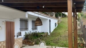 Eine überdachte Terrasse mit Tisch und Stühlen, eine Holz-Pergola und ein weißes Gebäude mit vergitterten Fenstern.