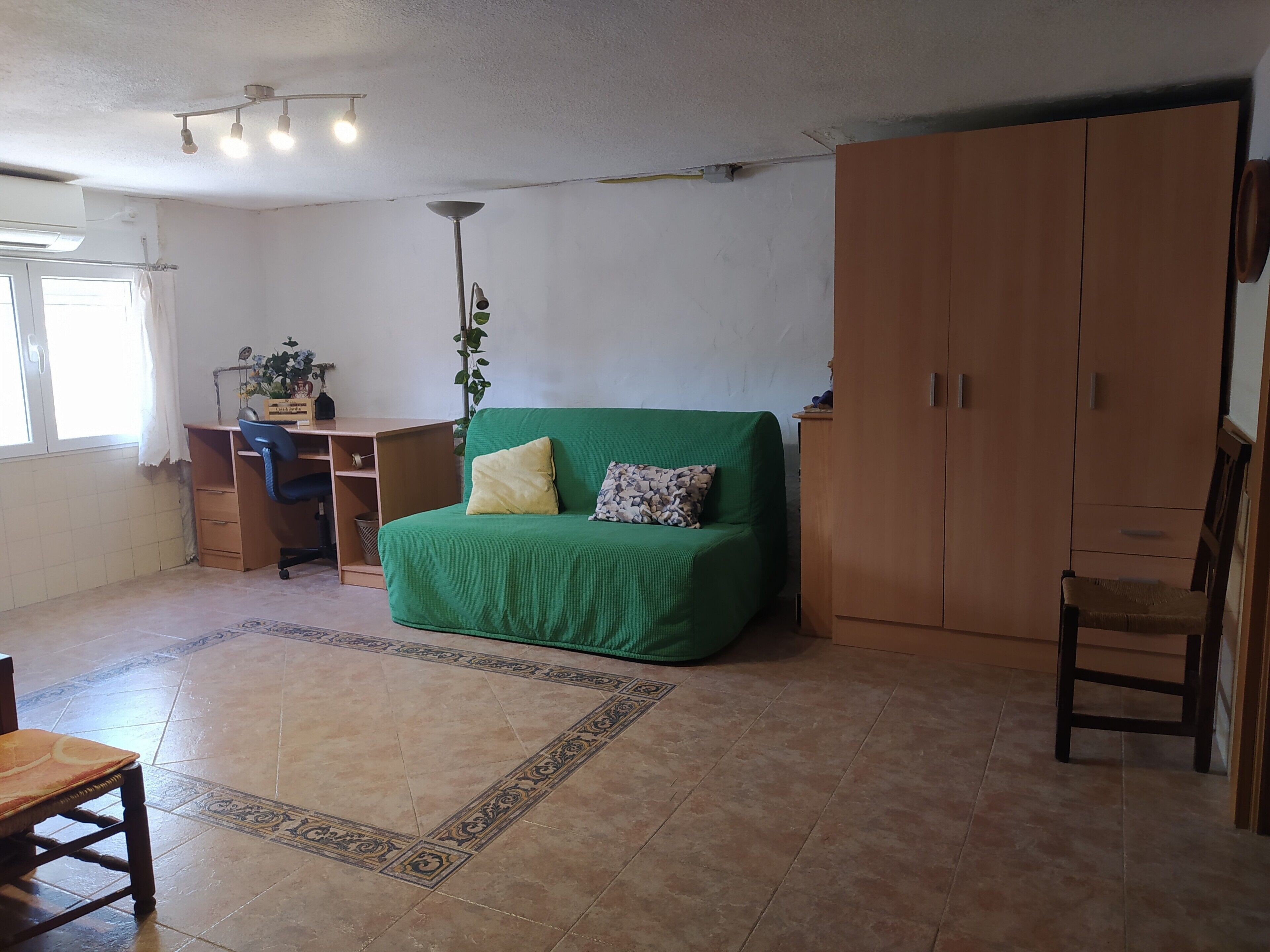 A room with a green sofa, wooden wardrobe, desk with chair, and a plant.