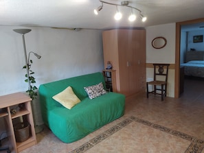 Un sofá verde con dos almohadas, un armario de madera, una mesita de madera, una lámpara de pie y una cama al fondo.