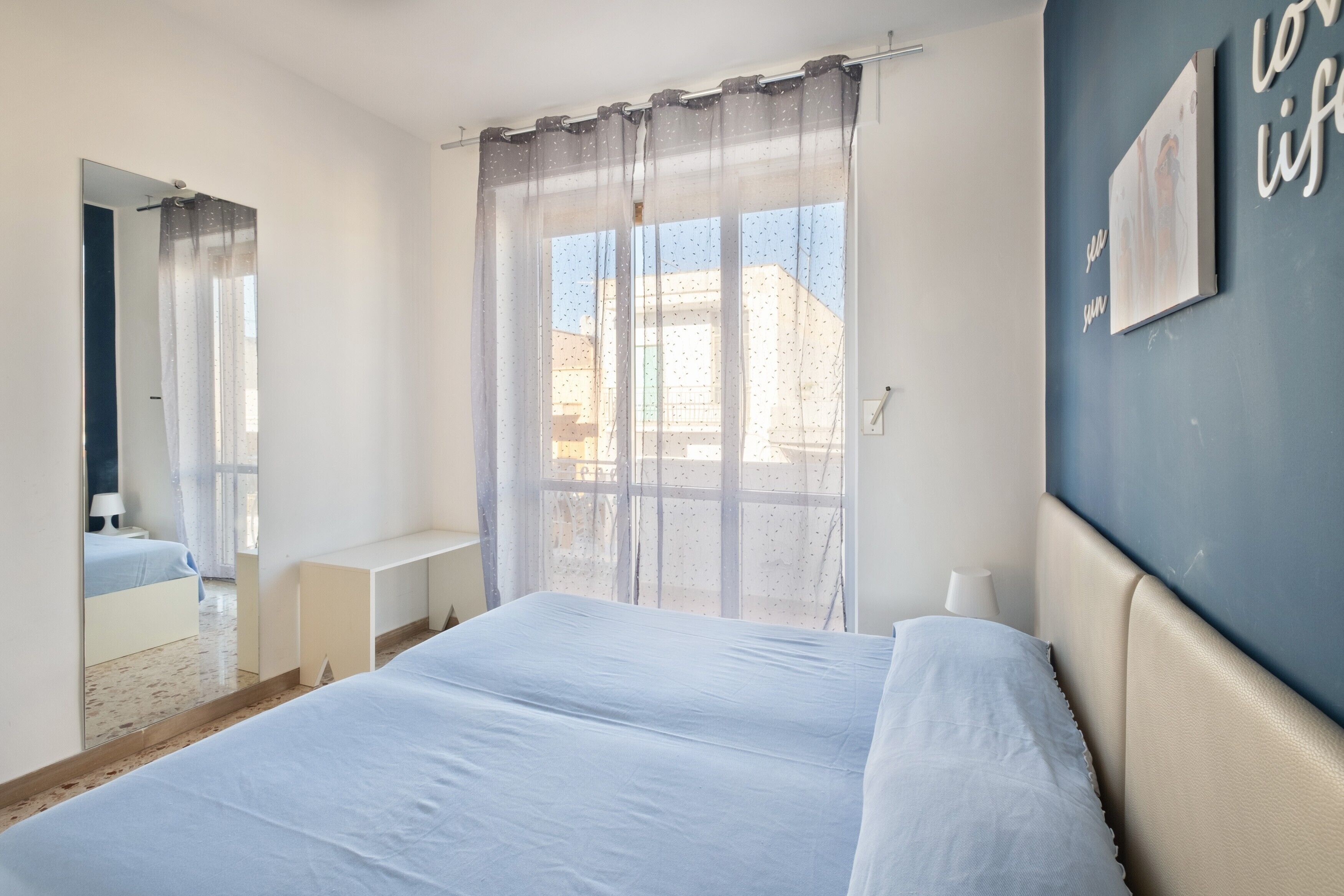 A bedroom with a bed, a window with sheer curtains, a bedside table, and a wall with a framed picture.