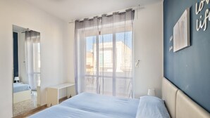A bedroom with a bed, a window with sheer curtains, a bedside table, and a wall with a framed picture.