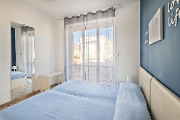 A bedroom with a bed, a window with sheer curtains, a bedside table, and a wall with a framed picture.