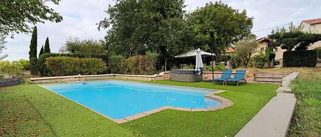 Ein rechteckiger Swimmingpool mit einer umliegenden Rasenfläche, einer Whirlpoolanlage und einer überdachten Sitzgelegenheit mit Stühlen.