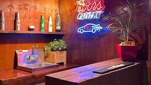 A bar area with a neon sign, wooden shelves with bottles, a sink, and a potted plant.
