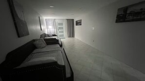A hotel room with a large bed, a smaller bed, and a sofa. There is a window with curtains, a door, and a wall-mounted picture.