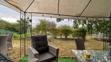 Een terras buiten met rieten meubilair, een glazen tafel en een overkapping.