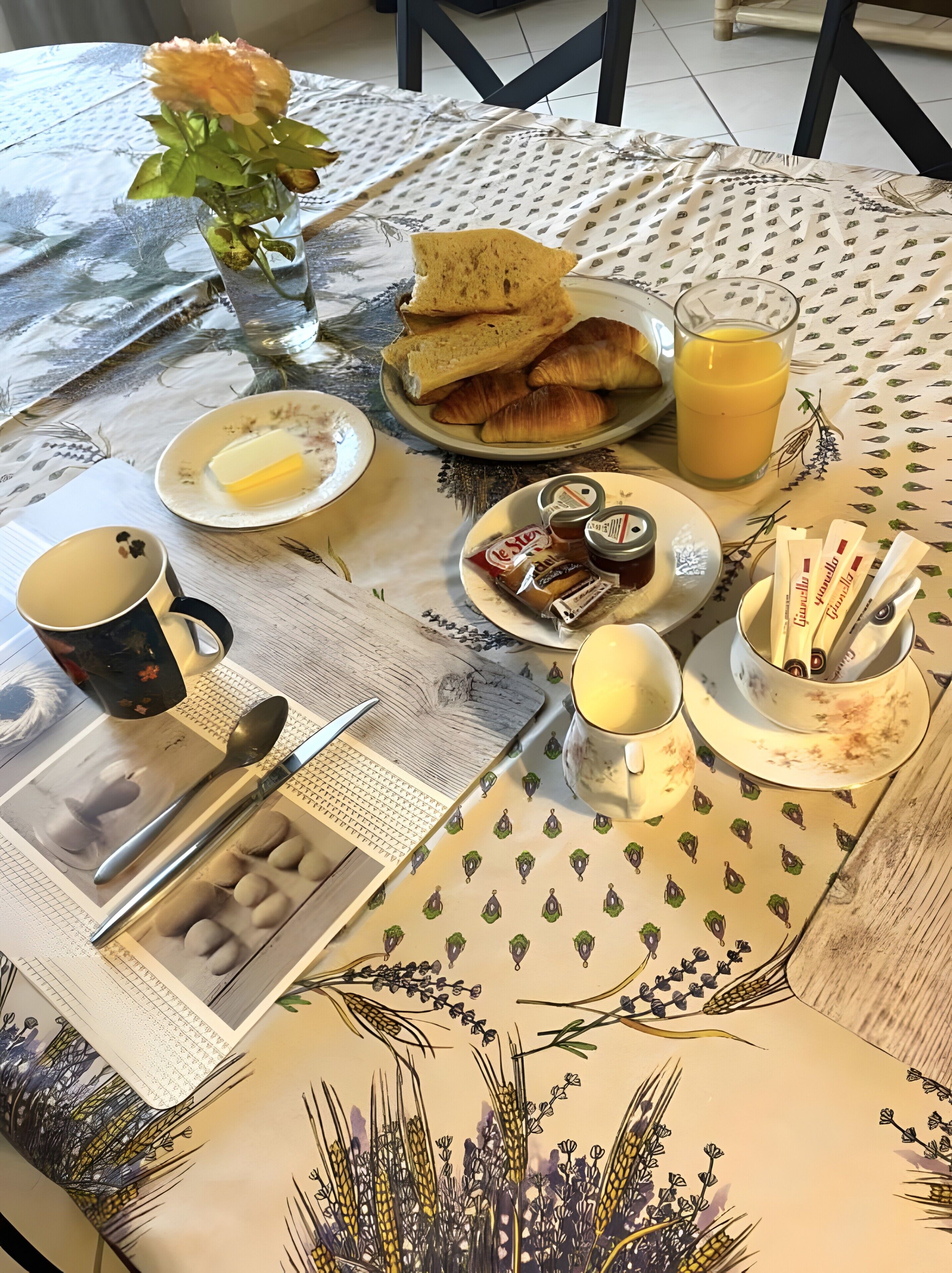 Una mesa de desayuno con cruasanes, mantequilla, mermelada, un vaso de zumo de naranja, una taza, una cuchara, un cuchillo y un periódico.
