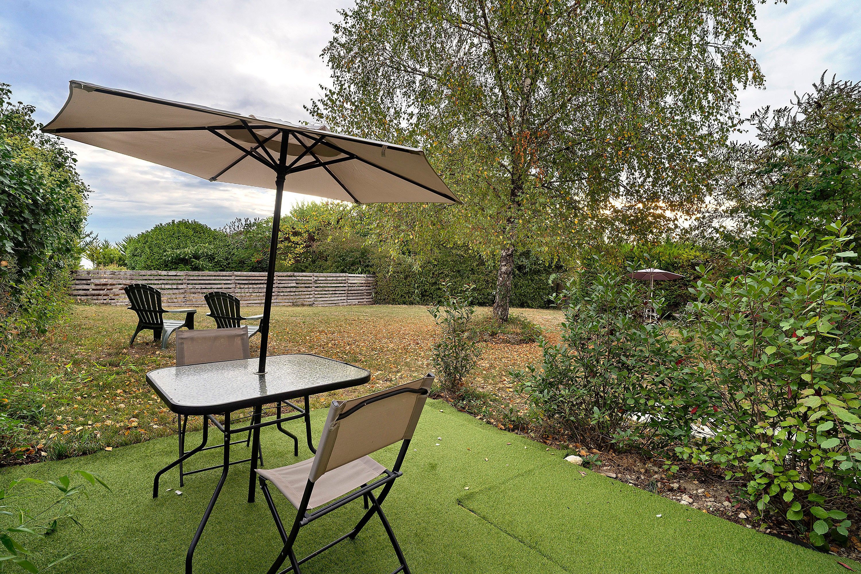 An outdoor patio area with a table and chairs, a large umbrella, and a grassy lawn.