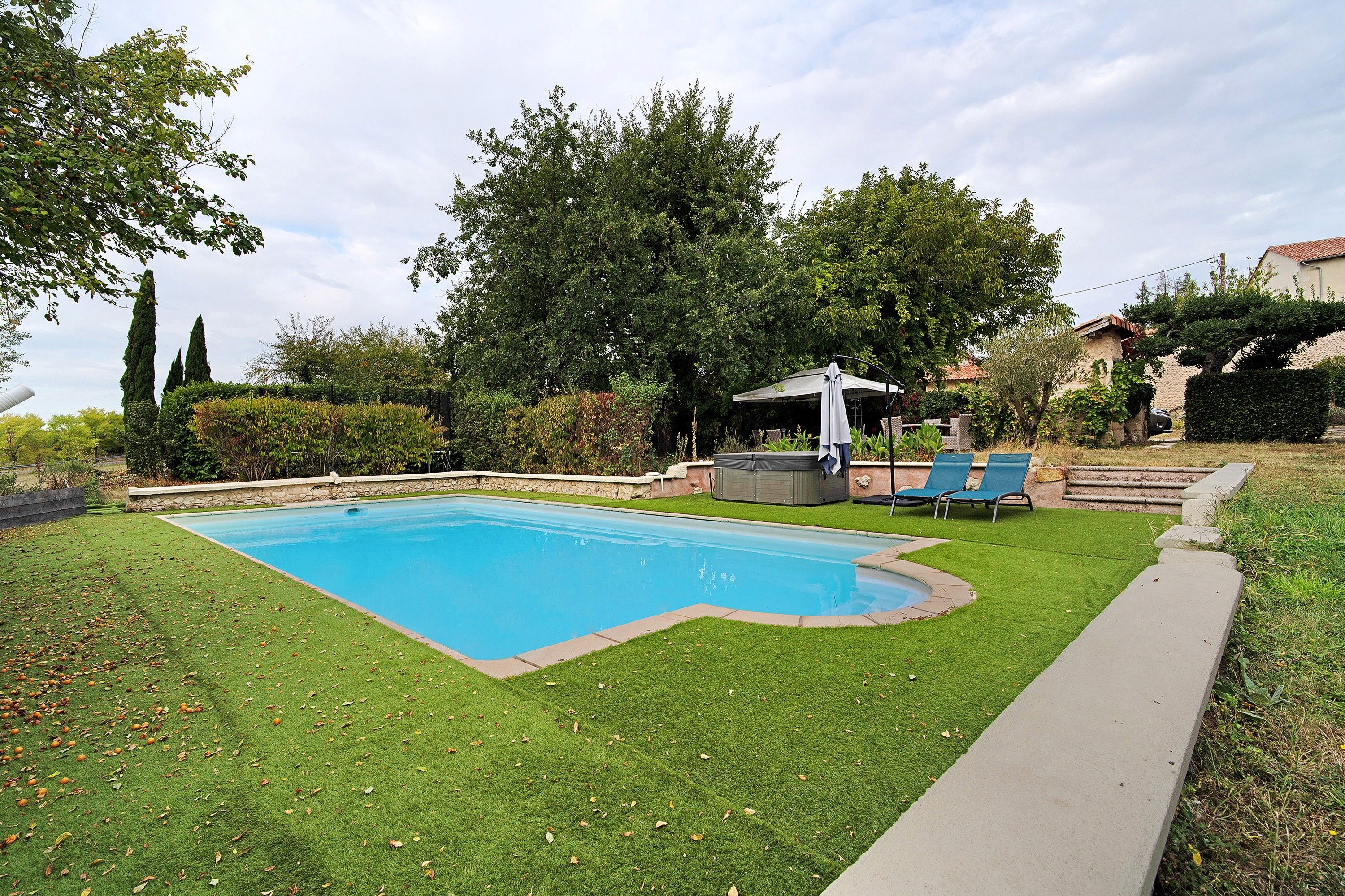 Una piscina rectangular con un área verde alrededor, un jacuzzi y una zona de estar techada con sillas.