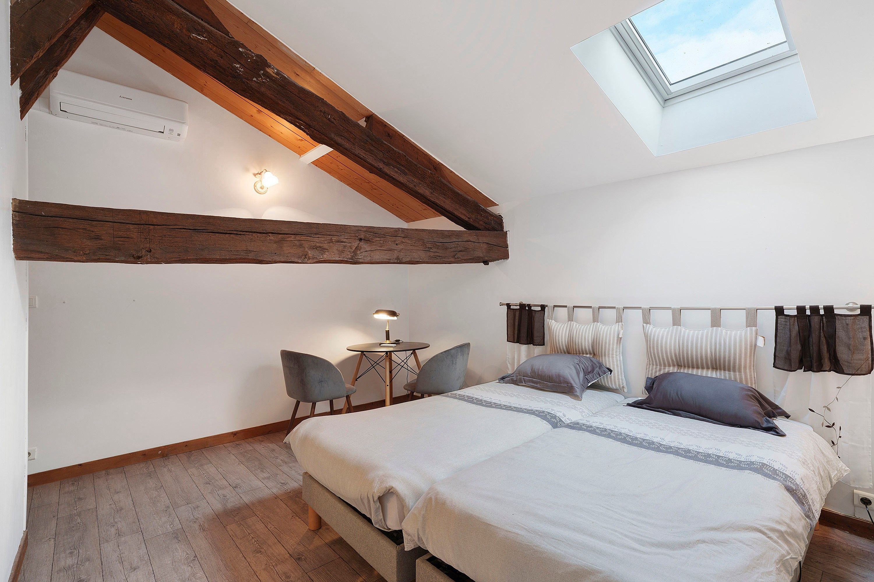 Un dormitorio con techo inclinado, tragaluces, una cama con almohadas, una mesita de noche y dos sillas.