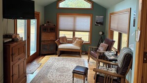 A cozy living room with a wooden ceiling, a large window with blinds, a television, a wooden cabinet, and a comfortable seating area with a rug and cushions. - Rustic Big Sky "Cabin" (Superior)