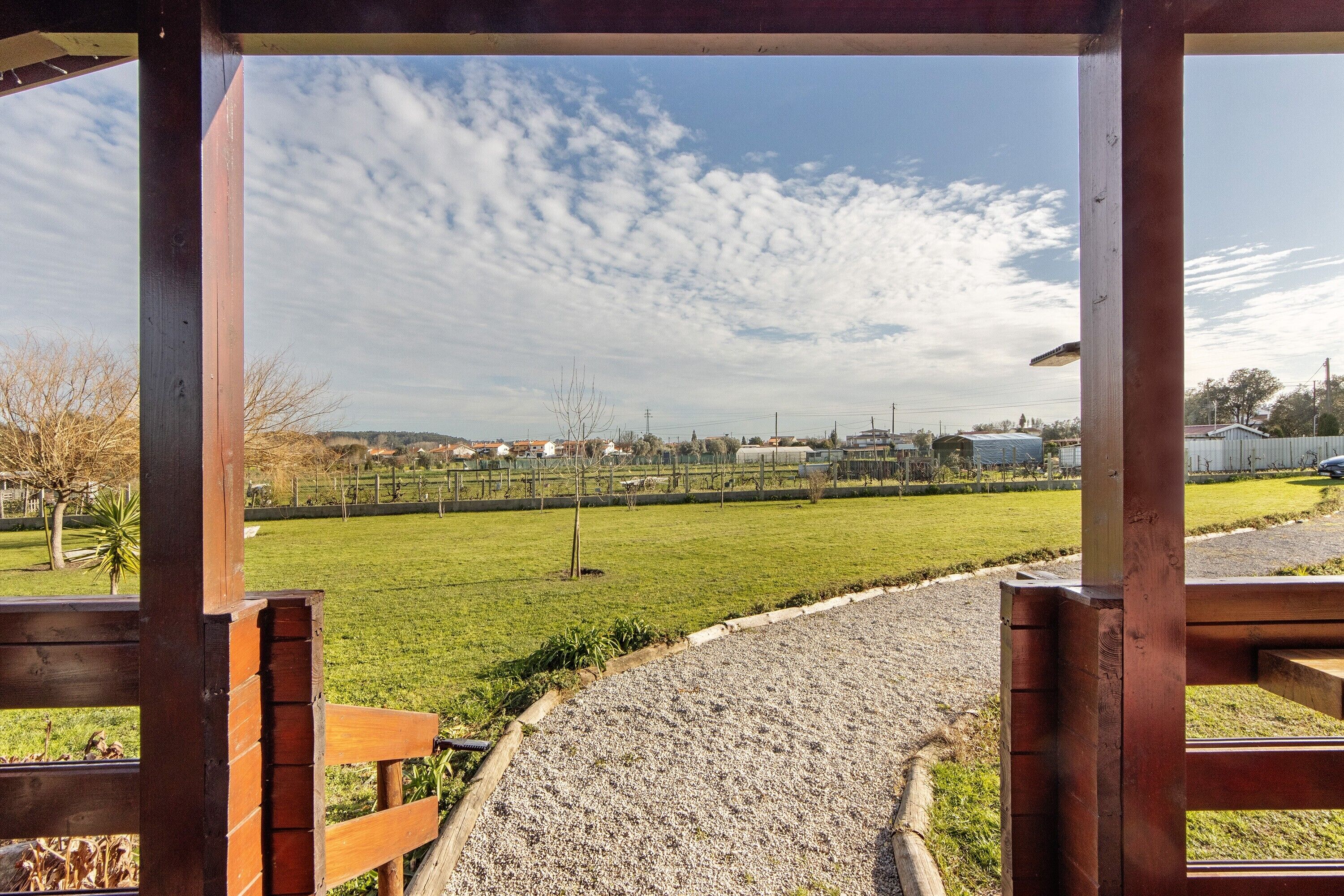 Uitzicht door een houten constructie, met een grindpad dat naar een groen veld en gebouwen in de verte leidt.