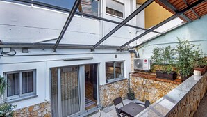 A residential building with a stone facade, a balcony with potted plants, and a sliding glass door leading to an interior space.
