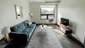 Una sala de estar moderna con un sofá azul, una mesa redonda, un televisor y un ventanal con persianas.