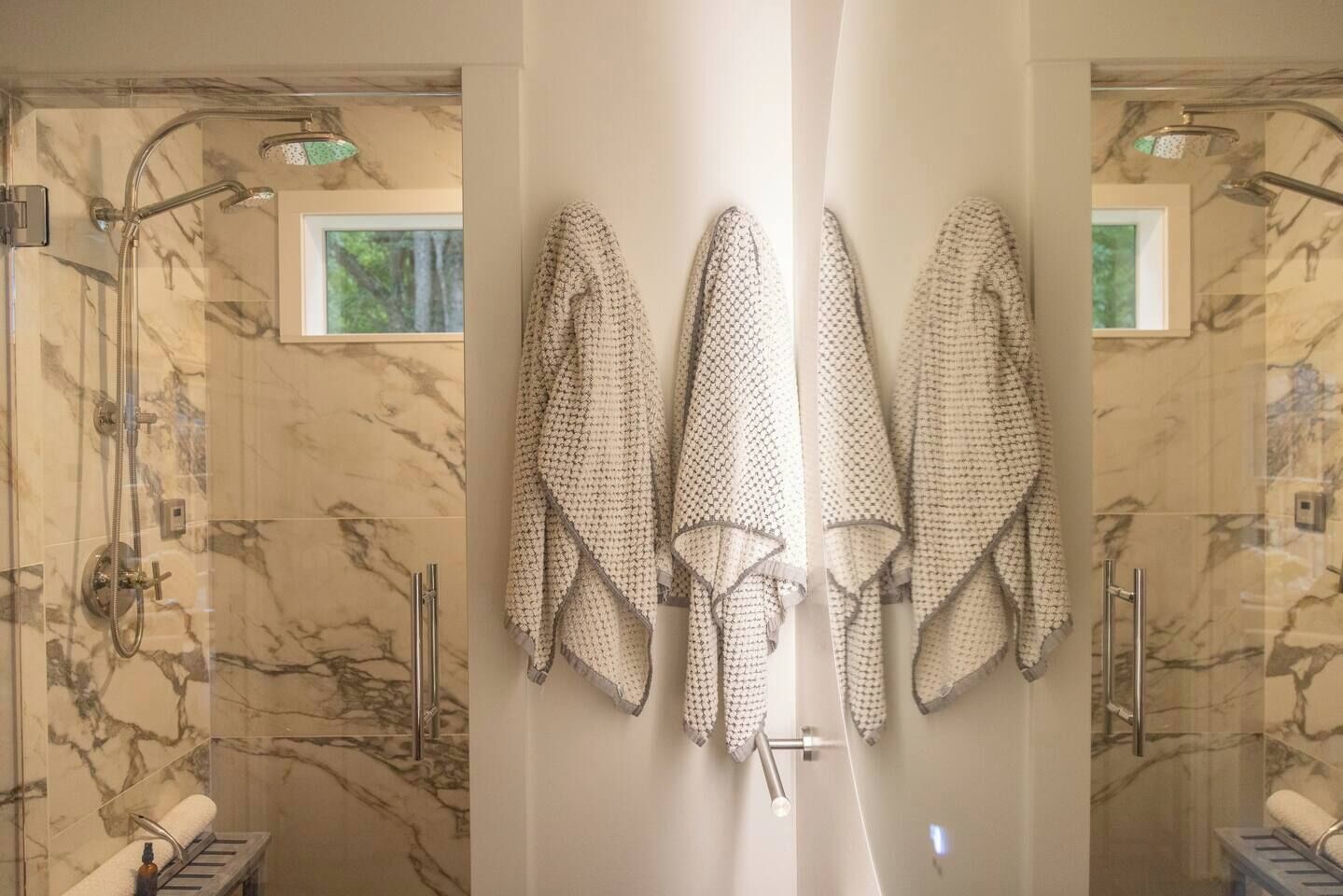 A bathroom with a shower, a towel rack with towels, and a window with a view of greenery.