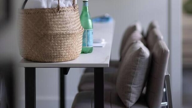 Una cesta de mimbre con una planta sobre una mesa, una botella verde y un cojín en una silla.