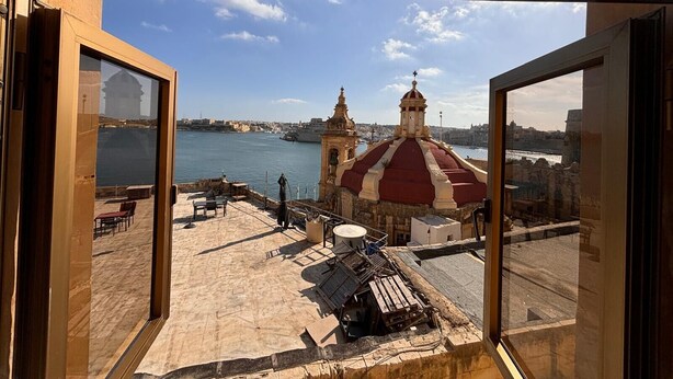 A view from a room with large windows overlooking a historic building with a red roof, a body of water, and a distant cityscape. - Valletta heart two, 3-room superior apartment (Valletta)