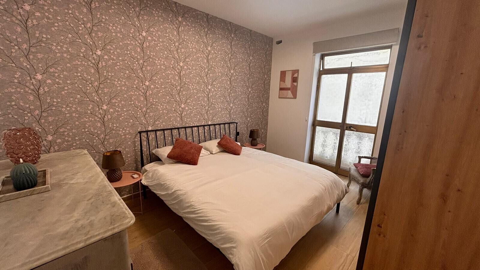 A bedroom with a bed, bedside tables, a nightstand, a window, and a wall with a floral pattern.