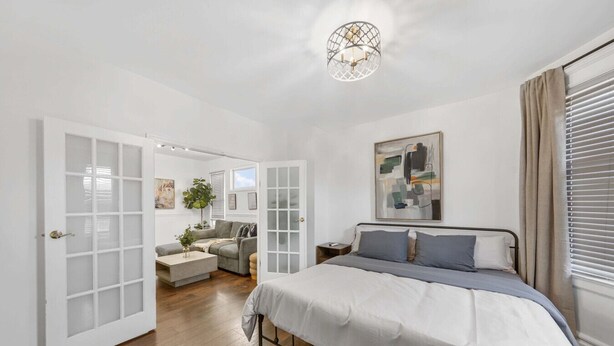 A bedroom with a bed, a nightstand, a window with blinds, and a door leading to another room.