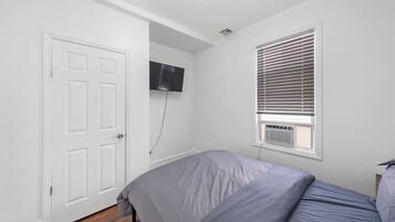 Une chambre à coucher avec un lit, une porte, un téléviseur fixé au mur et une fenêtre avec des stores.
