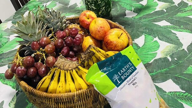 A table with a green patterned tablecloth, a wicker basket of fruits, a vase of pink flowers, and a bag of 'Earth's Boundry' snacks. - Charming 3-bedroom house with AC and WiFi in amazing Suva (Suva)