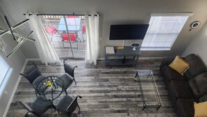 Una sala de estar con sofá, televisor, mesa y una silla. Se ve una ventana con persianas y vistas a muebles de exterior.