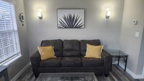Un salón con un sofá de cuero marrón oscuro, dos almohadas amarillas, una mesa de centro de cristal y una obra de arte abstracta colgada en la pared.