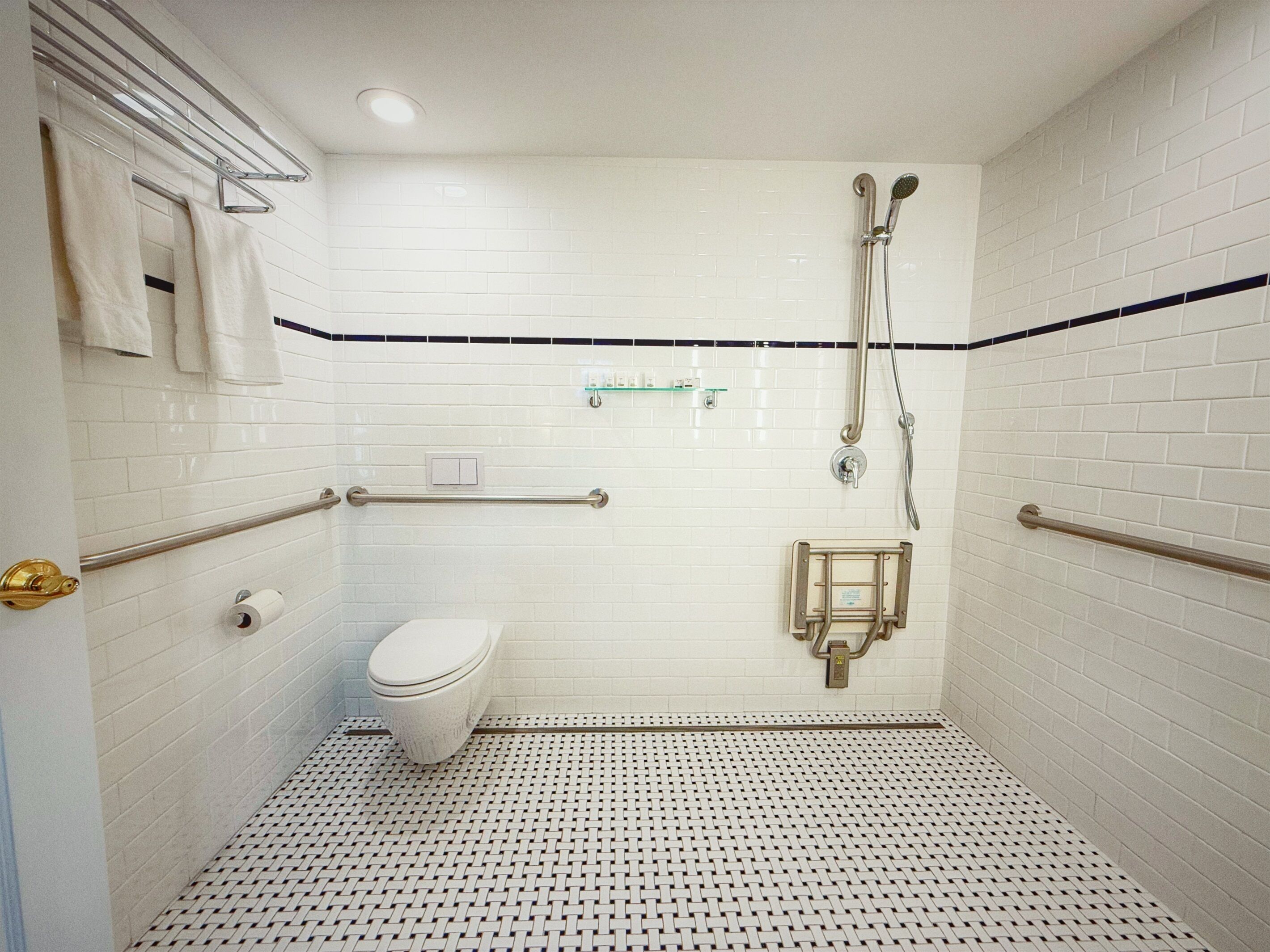A small, white-tiled bathroom with a toilet, a shower, and a handrail.